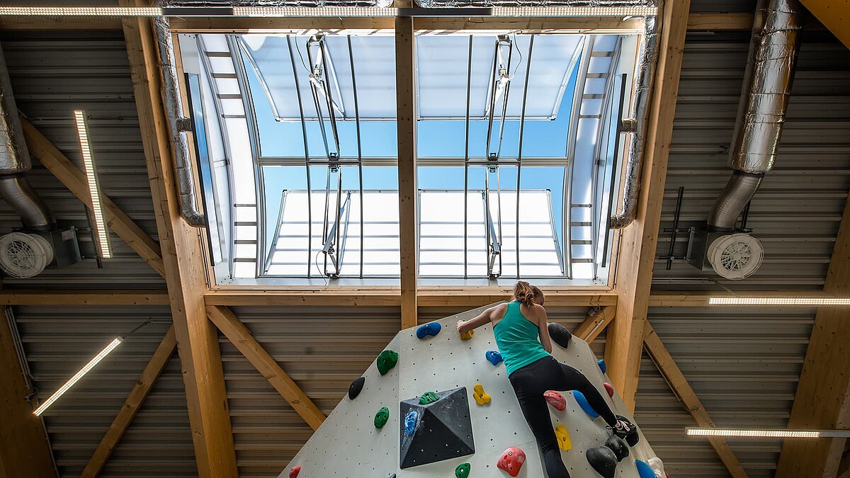 Kletterzentrum OWL mit Kletterwand und geöffneter Lichtkuppel, durch die Tageslicht in die Halle fällt. Eine Frau klettert die bunte Wand hoch.