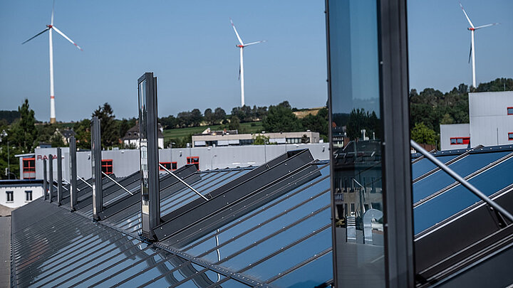 Dachfläche mit mehreren Glasdachelementen, Metallkonstruktion und Windrädern im Hintergrund.