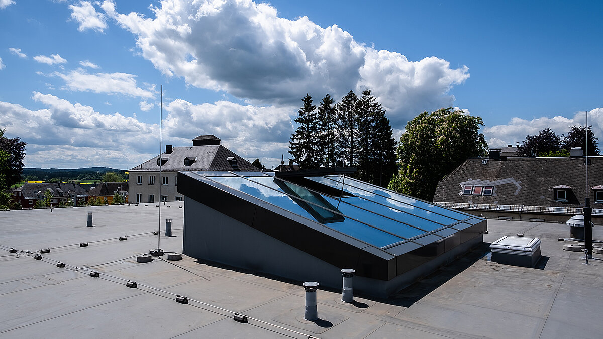 Schräges Glasdach auf einem Flachdach, moderne Dachverglasung mit geöffnetem Lüftungsflügel unter blauem Himmel