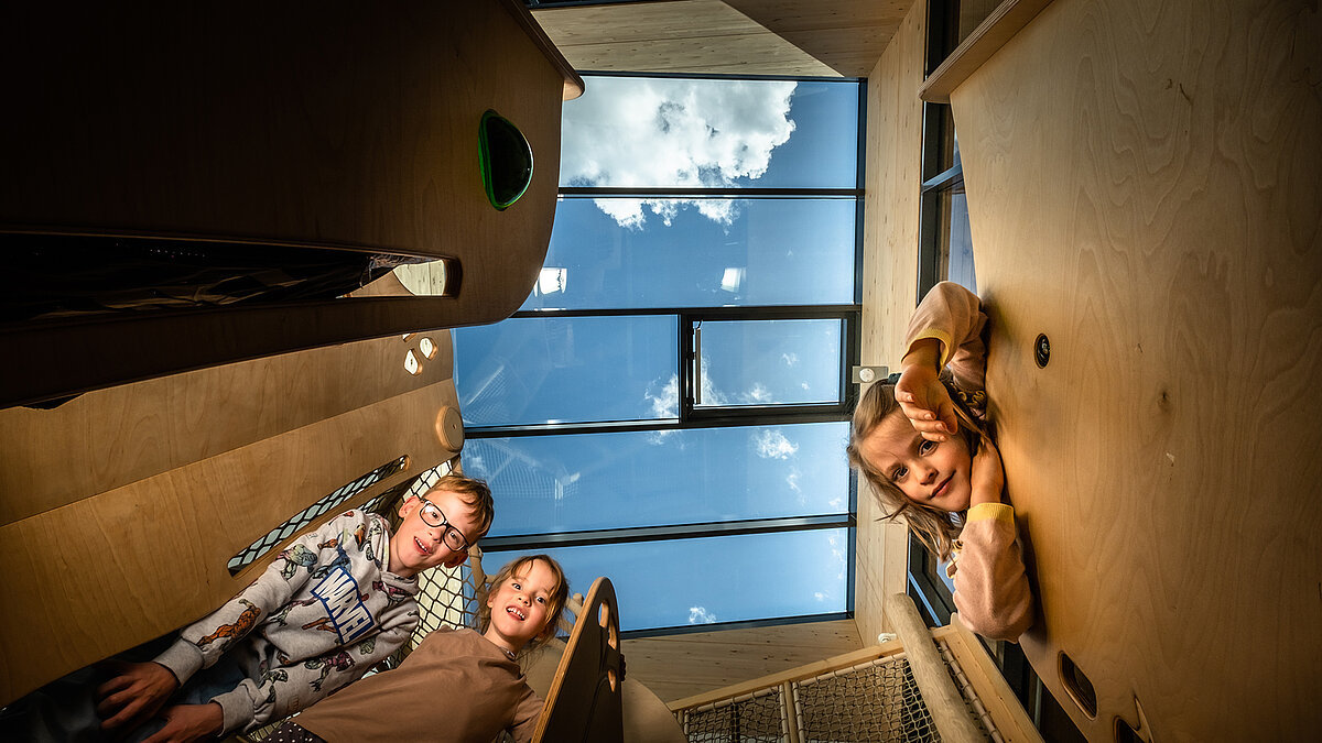 Kinder schauen nach unten in die Kamera, über ihnen ein großes Glasdach mit Blick in den Himmel, heller Innenraum mit Holzverkleidung