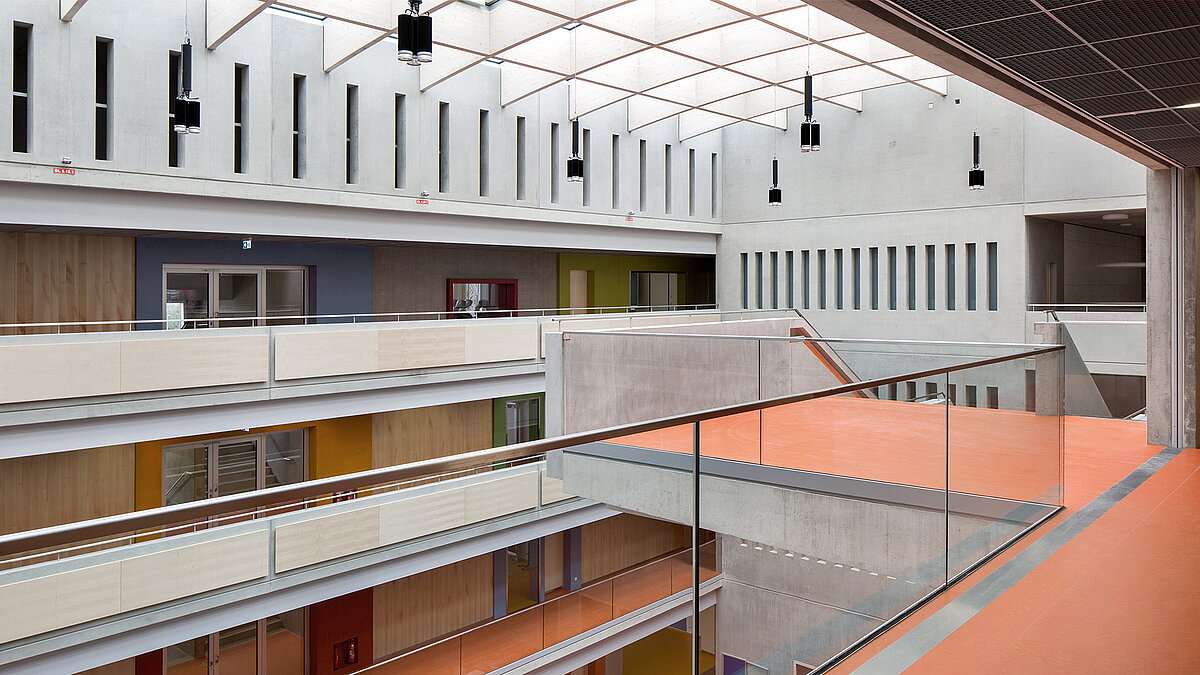 Modernes Atrium im Willibald-Gluck-Gymnasium in Neumarkt mit LAMILUX Glasdach, Betonwänden und farbigen Böden.