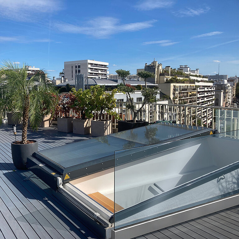 Moderne Dachterrasse mit geöffnetem Flachdach Ausstieg, Palmen, Pflanzkübeln und Stadtblick bei blauem Himmel