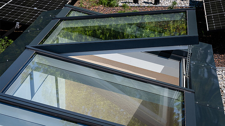 Geöffnetes Aluminium-Lichtband auf Flachdach mit Solarpanelen, Kiesbett und extensiver Dachbegrünung im Hintergrund