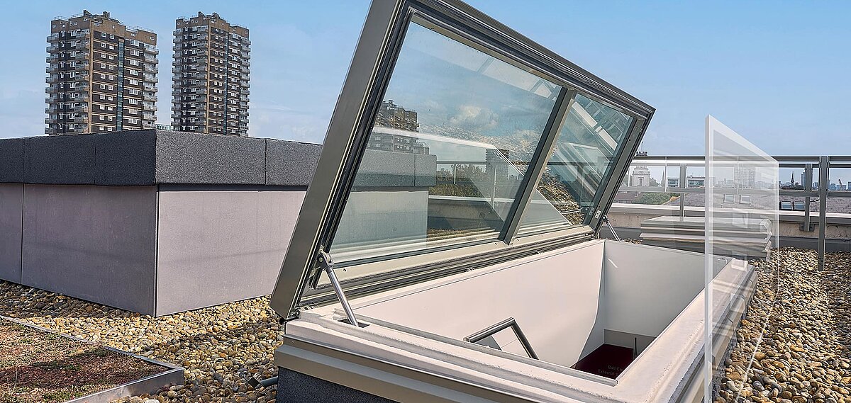 Geöffneter LAMILUX Flachdach Ausstieg auf einem begrünten und mit Kies belegten Dach, im Hintergrund Hochhäuser und Skyline von London.