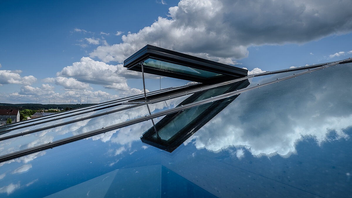 Detailansicht eines geneigten Glasdachs mit geöffnetem Lüftungsflügel mit Spiegelung von Wolken im Glas