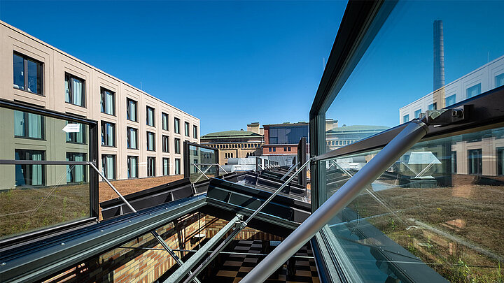 Geöffnetes Glasdach mit Metallrahmen, Blick auf begrüntes Flachdach und moderne Bürogebäude unter blauem Himmel