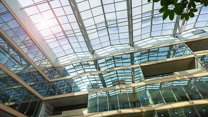 Modernes Gebäude-Atrium mit großem Glasdach, mehrstöckige Glasfassaden, Sonnenlicht und grüne Pflanze