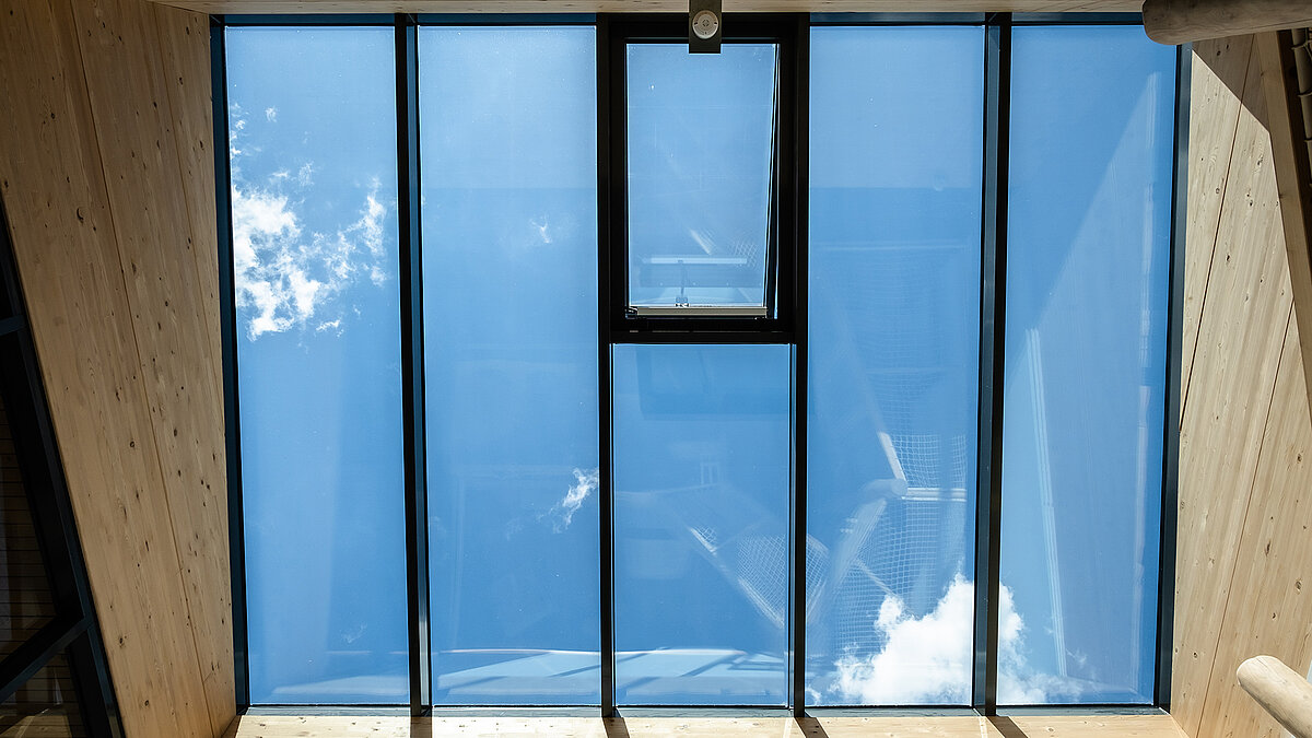 Blick durch ein großflächiges Glasdach mit schmalem Lüftungsflügel, blauer Himmel und Wolken über einem holzverkleideten Innenraum