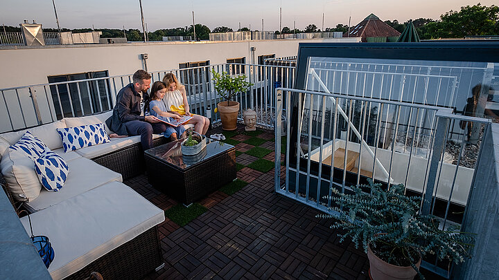 Familie sitzt auf einer gemütlichen Dachterrasse mit Lounge-Möbeln, Pflanzen und Abendstimmung.